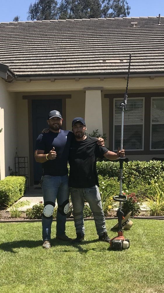 Two men stand on a lawn, one holding a weed whacker, in front of a house.