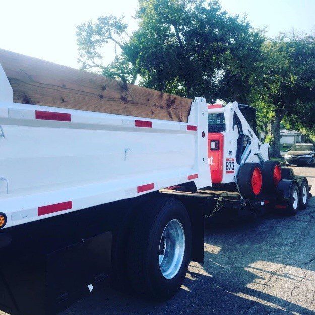 White dump truck with a Bobcat, carrying lumber. Red and white accents, sunny outdoor setting.