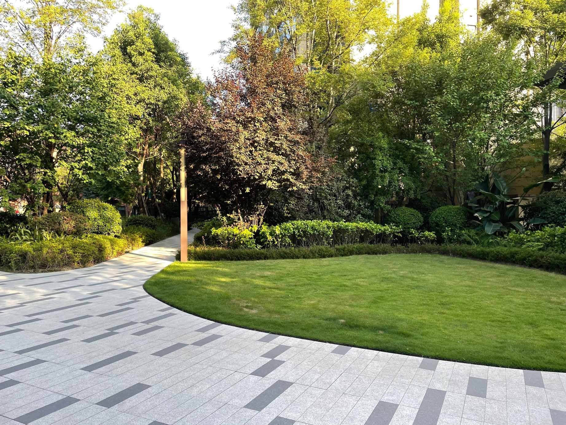 Lush green garden with paved walkway and grass lawn surrounded by trees and shrubs in sunlight.