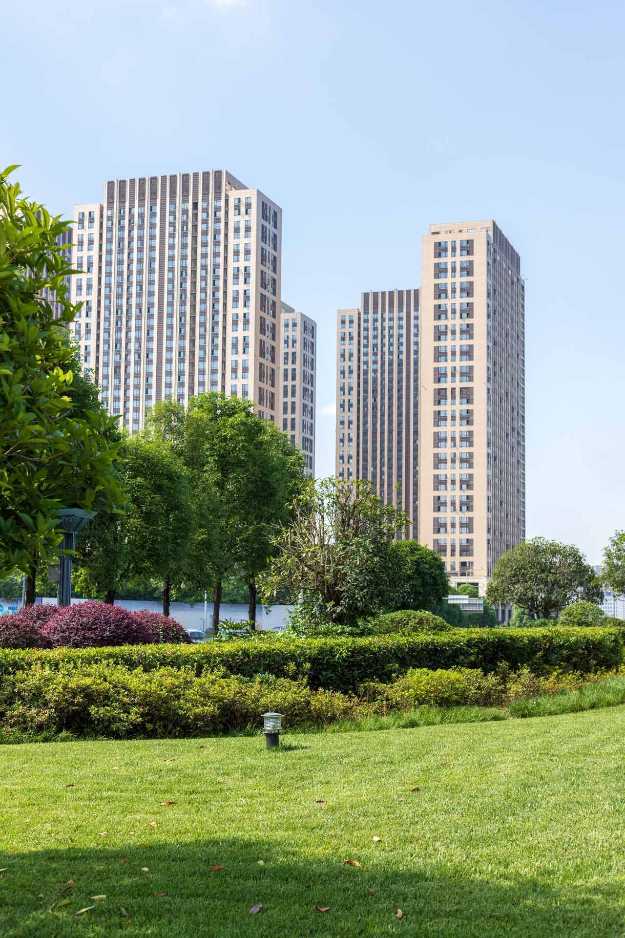 Tall beige buildings with many windows stand behind a green lawn with trees and bushes under a blue sky.
