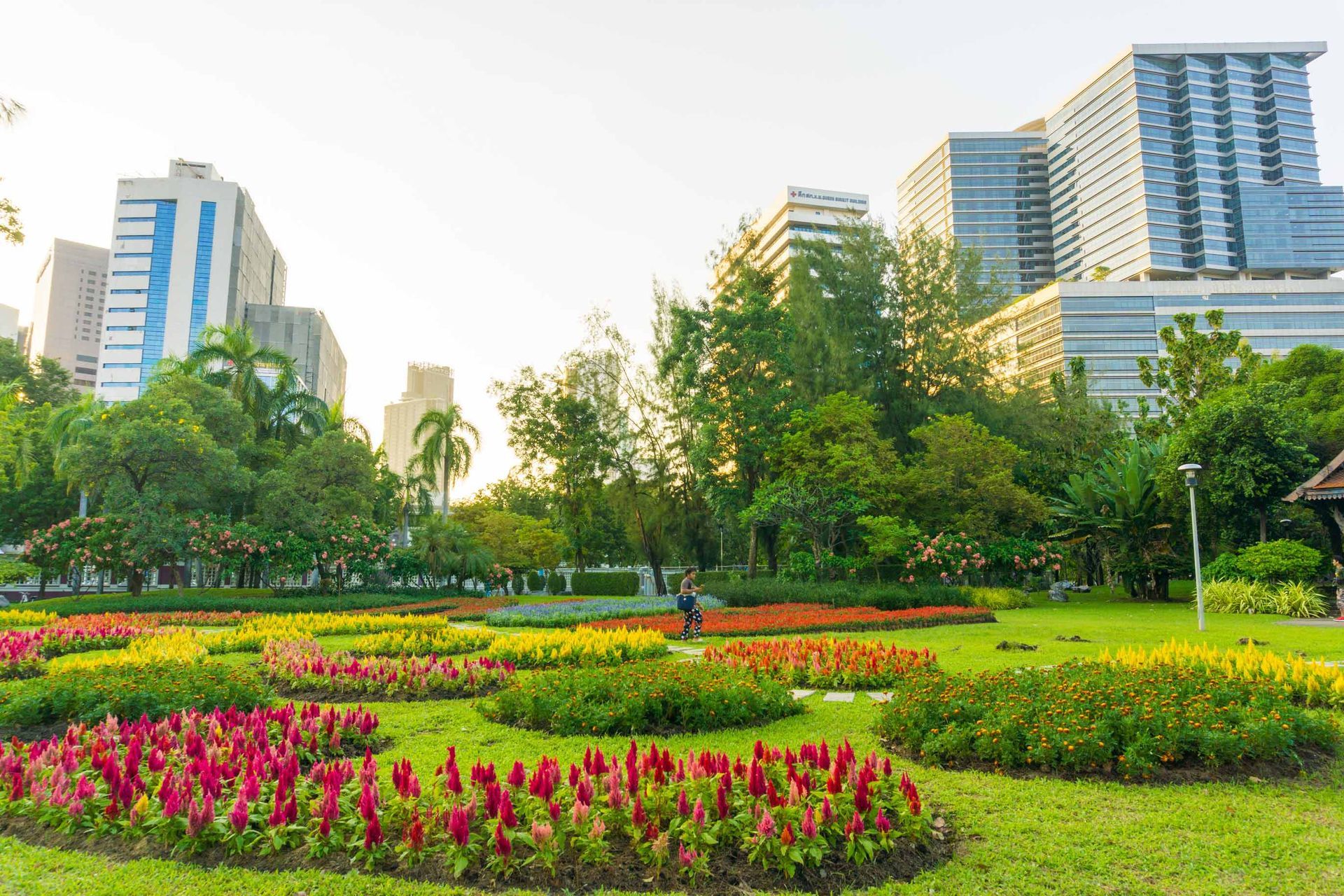 Colorful flowerbeds in a park with tall buildings in the background under a cloudy sky.