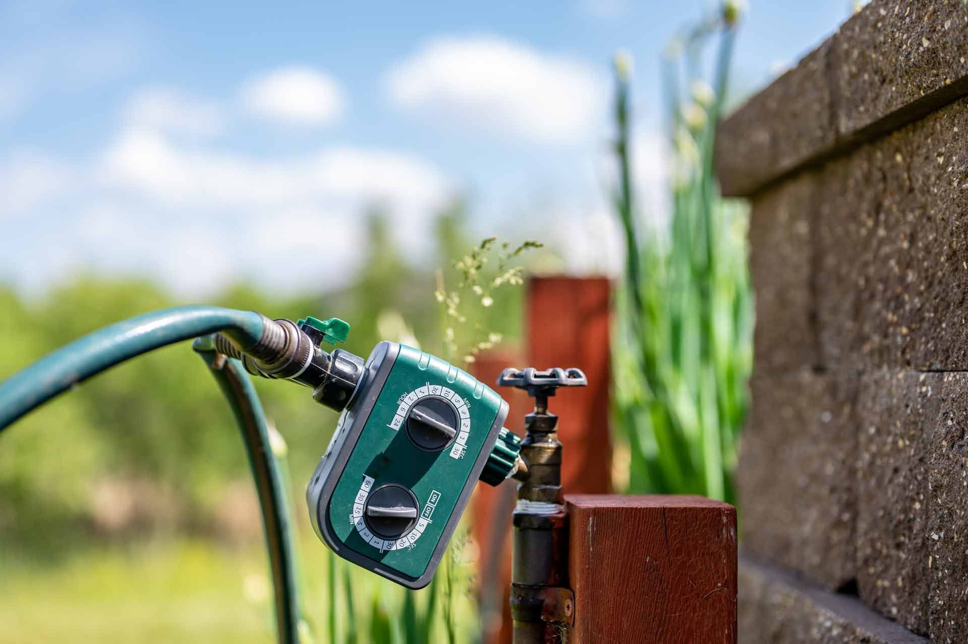 A green hose timer attached to a faucet sprays water outdoors near a brick wall and plants.