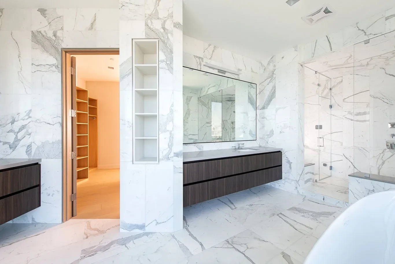 A bathroom with marble walls , two sinks , a tub and a walk in closet.