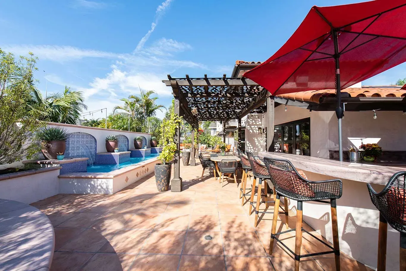 A patio with a bar , chairs , umbrellas and a pool.
