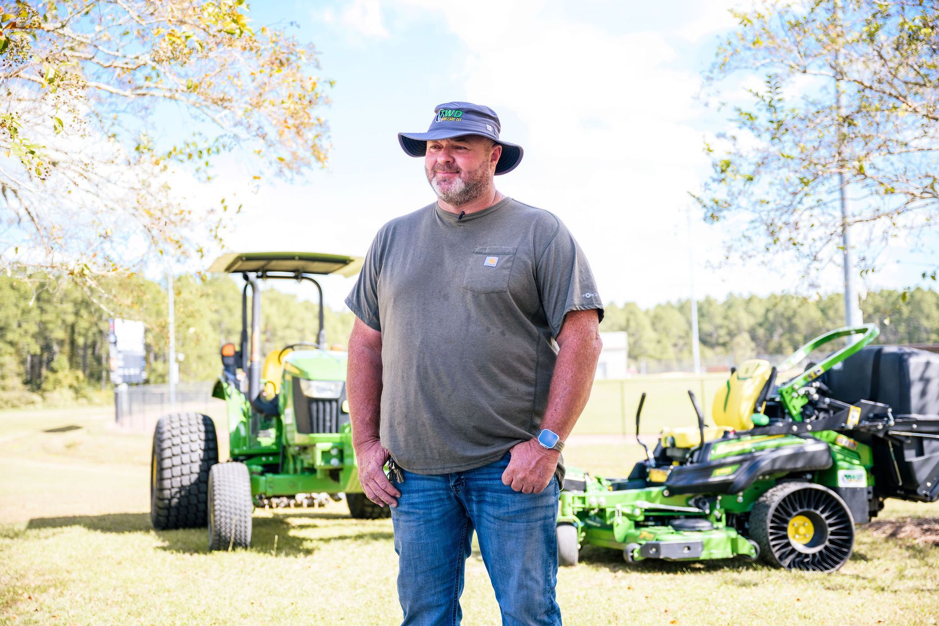 A man in a hat is standing in front of a green tractor and mower.