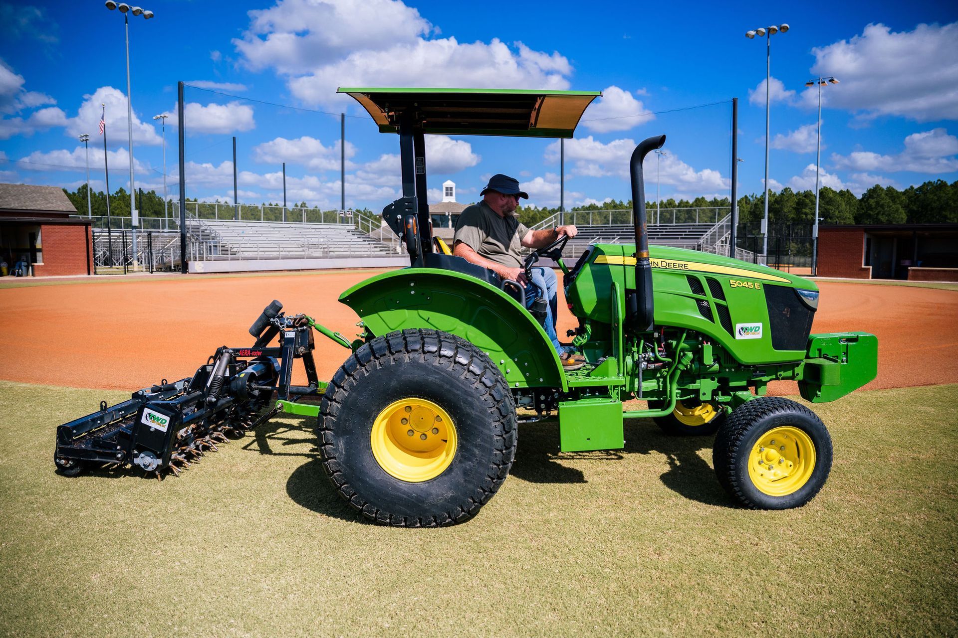 A man is driving a green utility tractor on a baseball field.