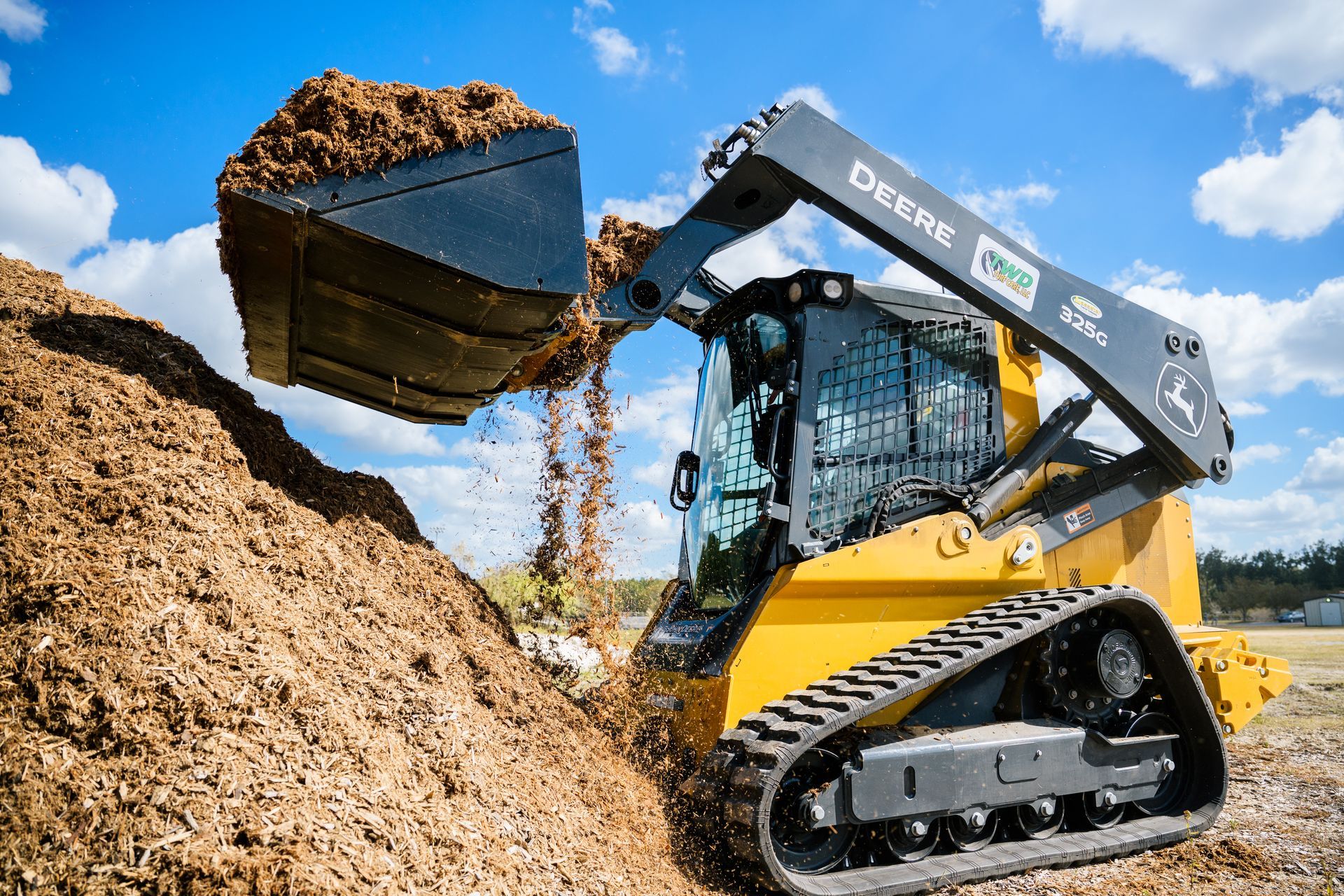 A compact track loader is loading dirt into a pile of wood chips.