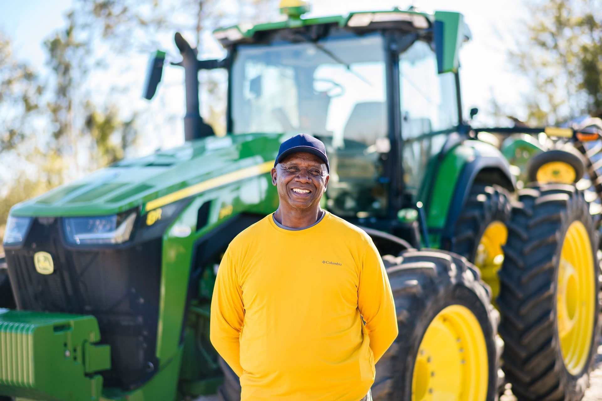 A man in a yellow shirt is standing in front of a green john deere tractor.