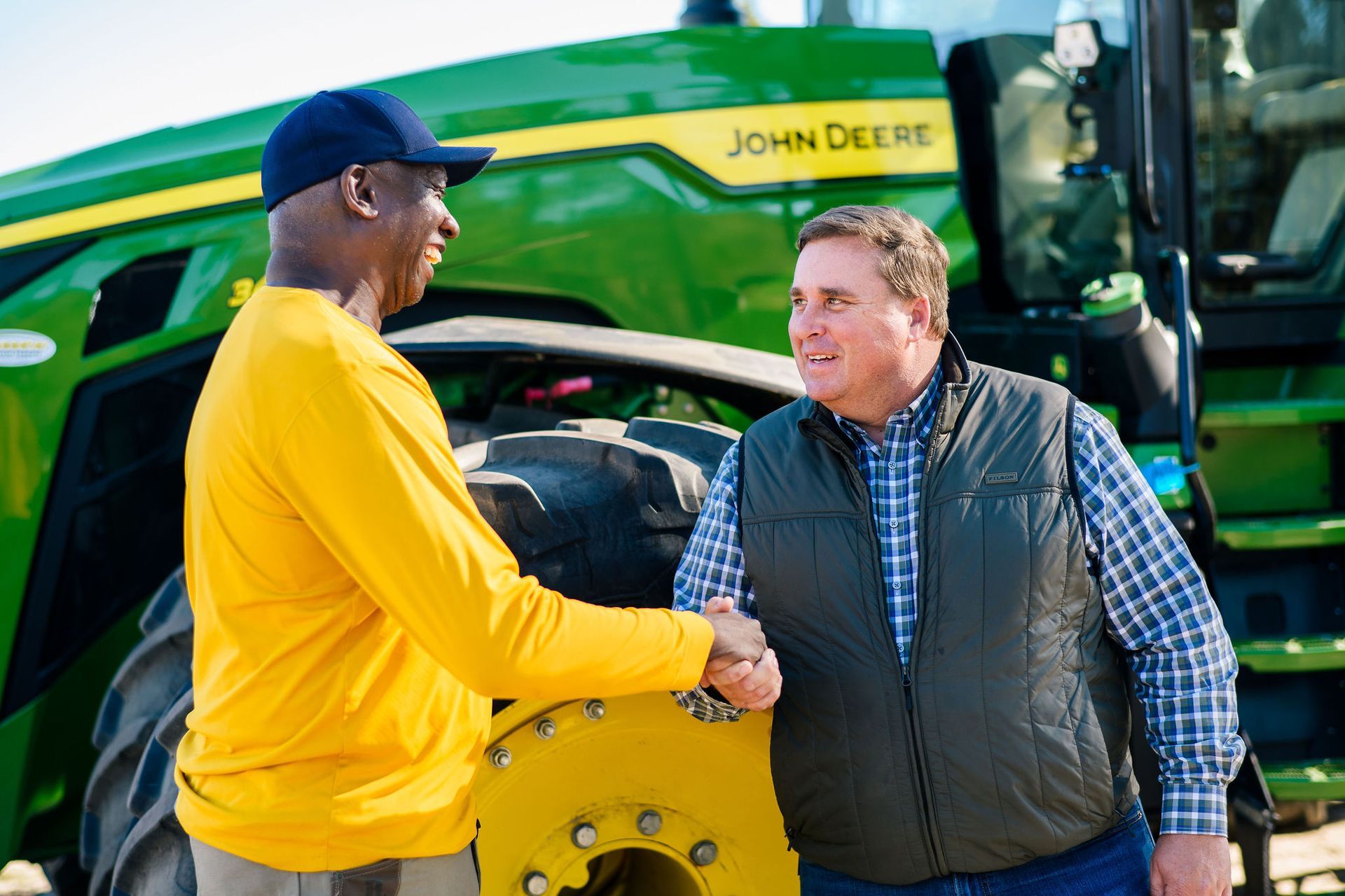 Two men are shaking hands in front of a john deere tractor.
