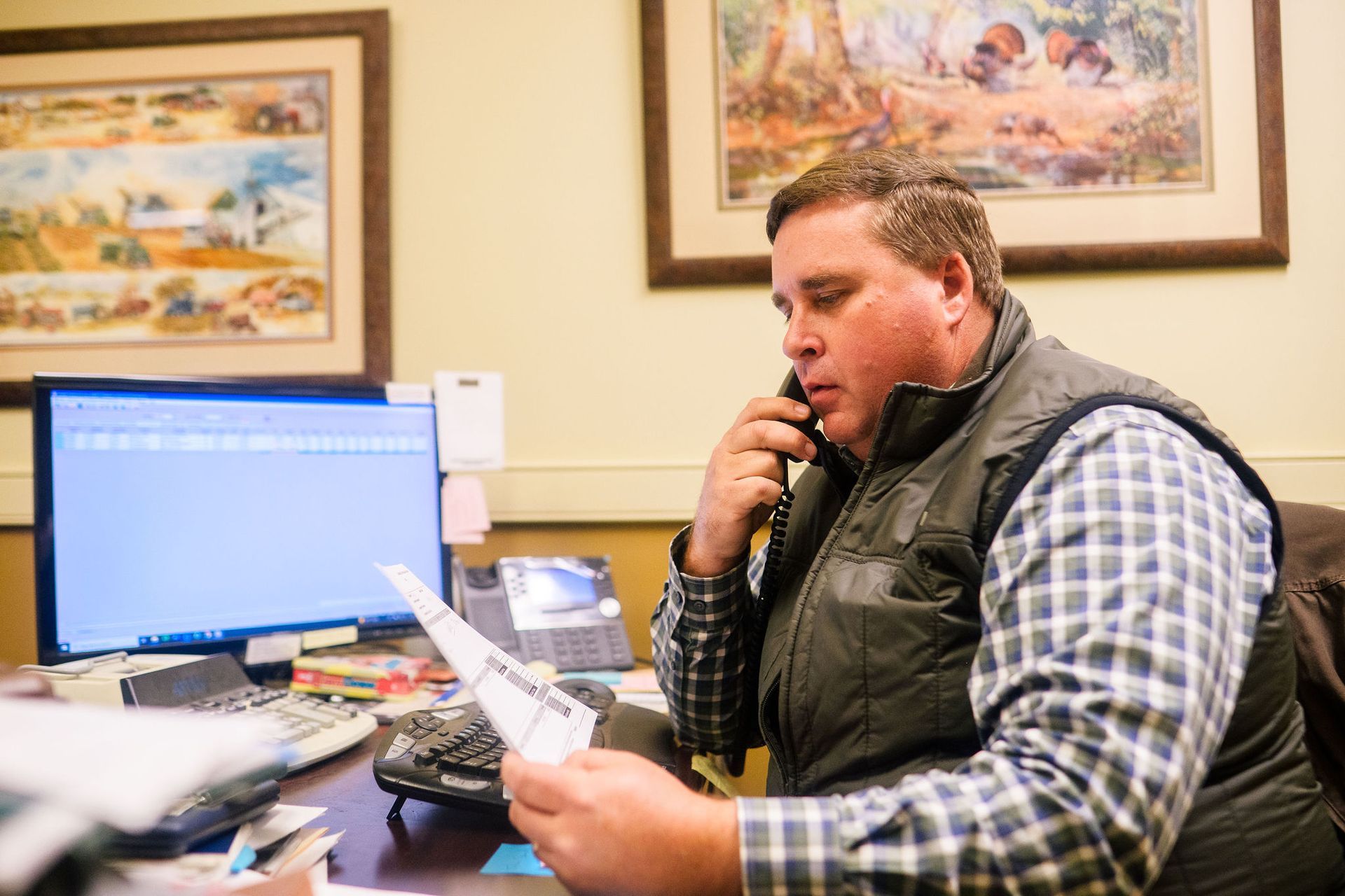 A man is sitting at a desk talking on a phone while holding a piece of paper.
