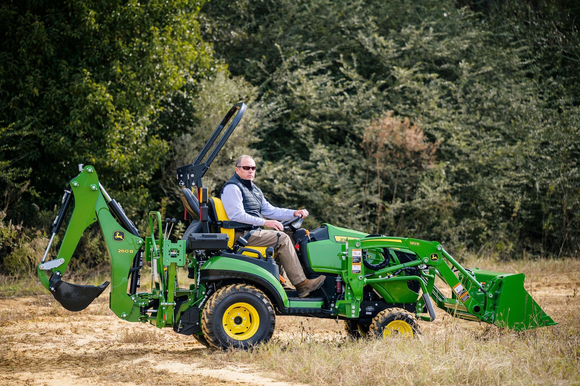 A man is driving a green tractor with a backhoe in a field.