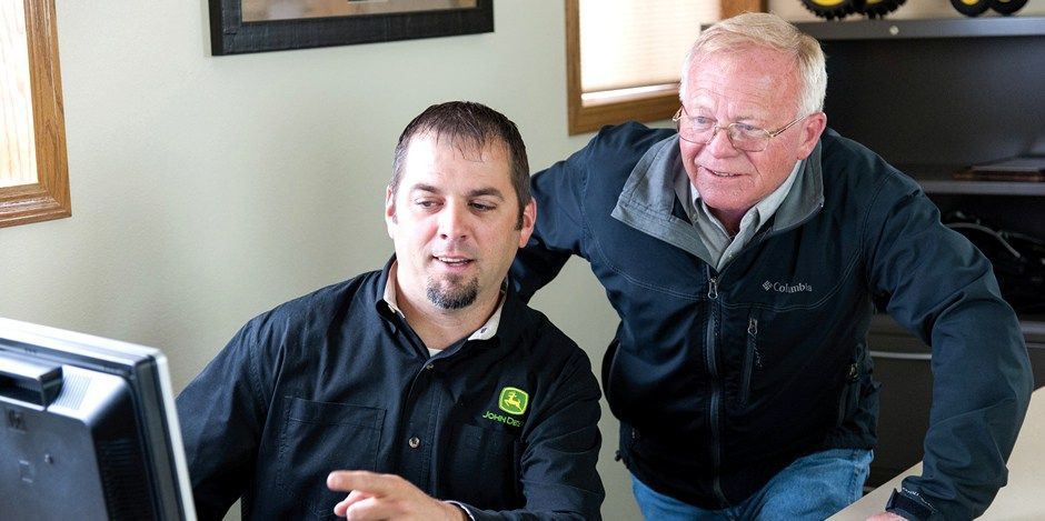 Two men in john deere jackets are looking at a computer screen.