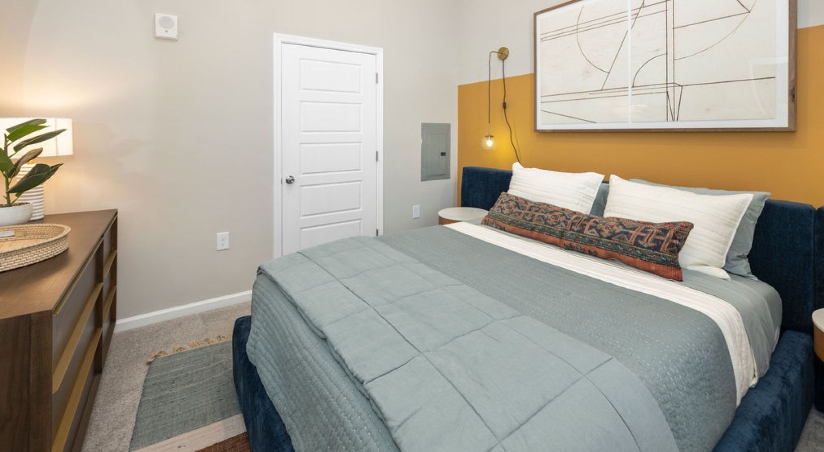 Bedroom in apartment with a large bed, blue upholstered headboard, gray bedding, and a dresser.