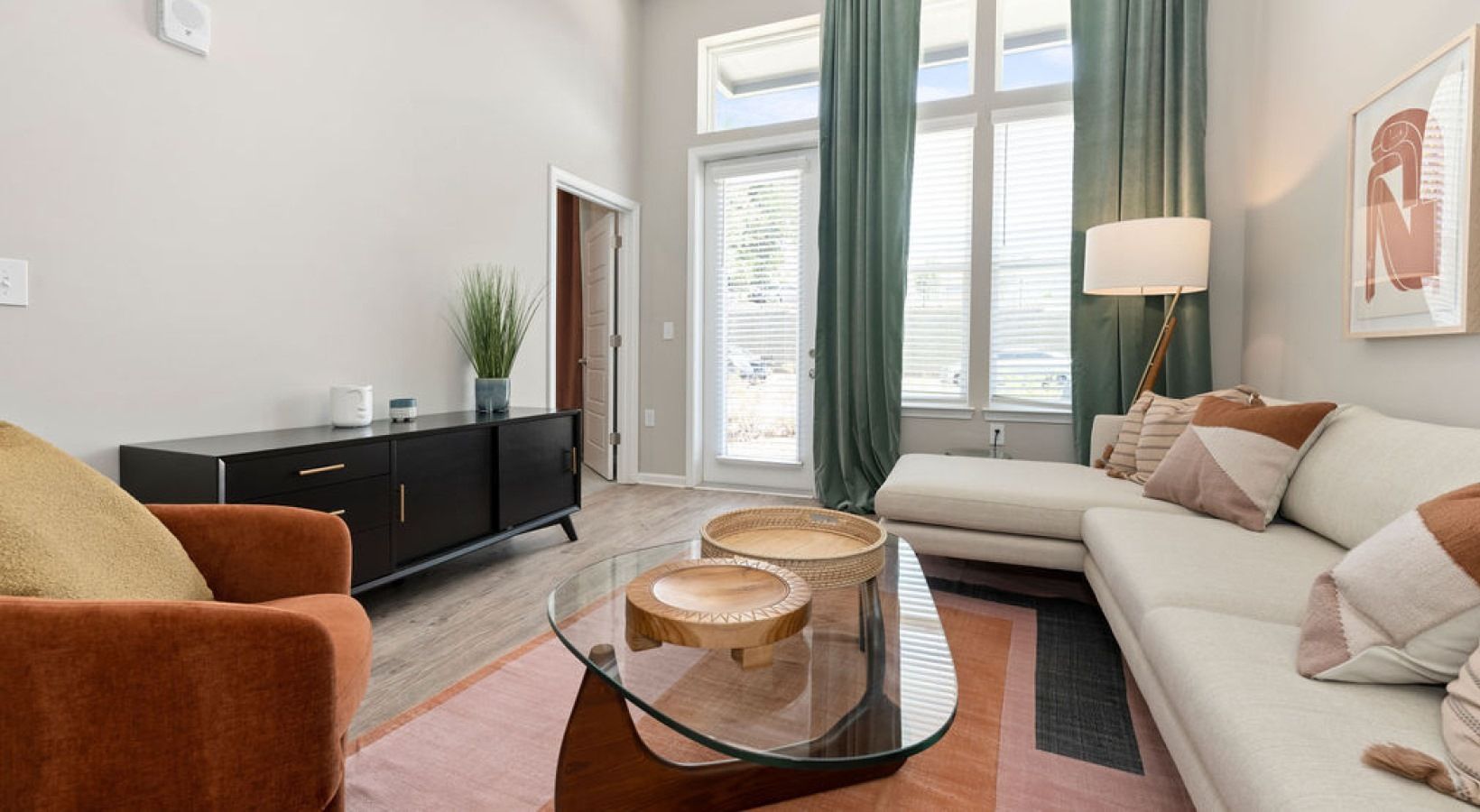 Living room in a modern apartment with a white sectional sofa, orange chair, glass coffee table, and tall windows with green curtains.