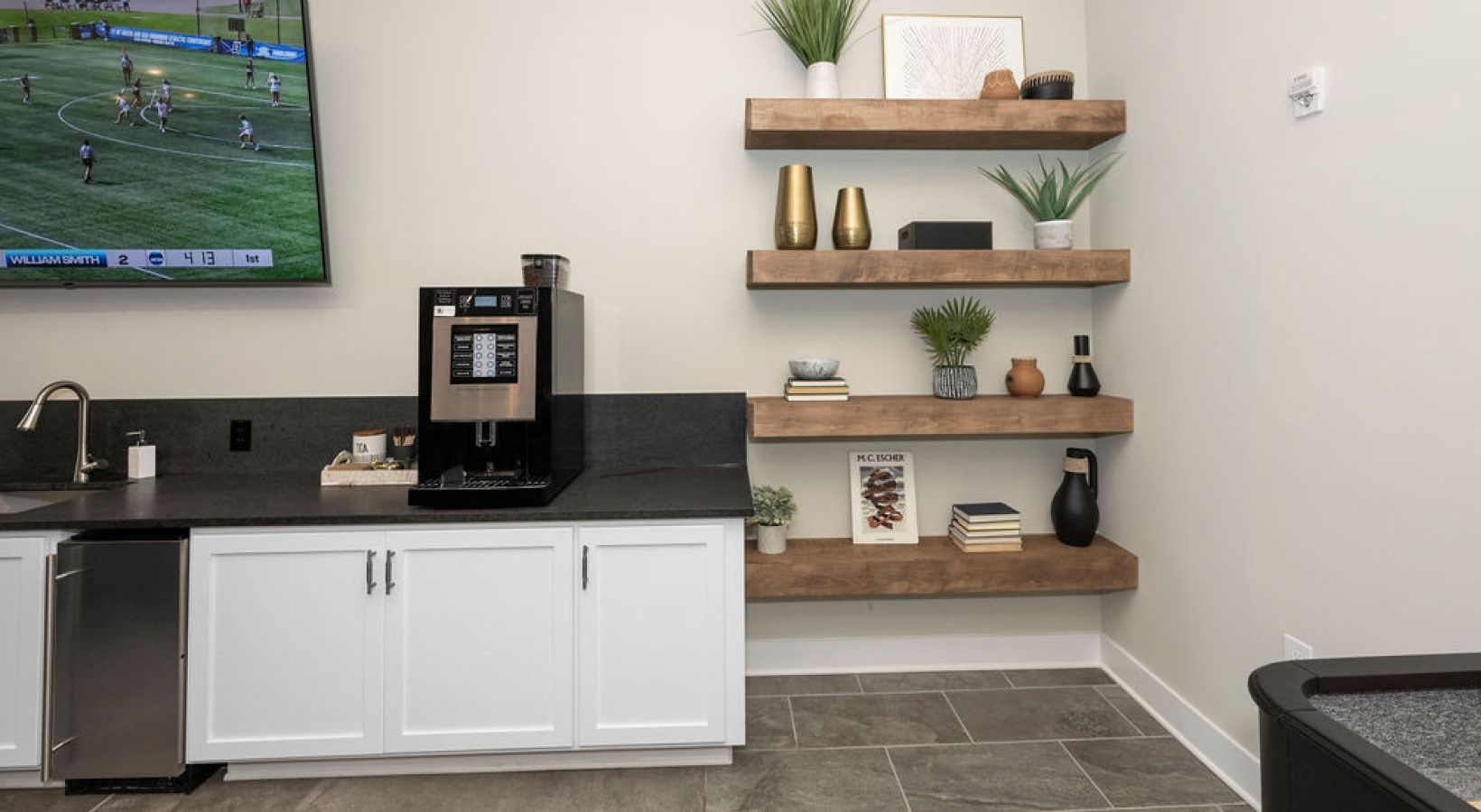 Modern community kitchenette with coffee station, white cabinets, and floating wood shelves.