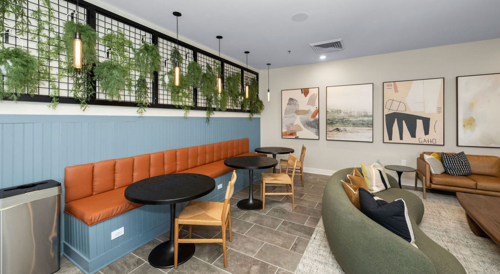 Interior community lounge with blue paneled wall, orange bench seating, plants, and abstract wall art.