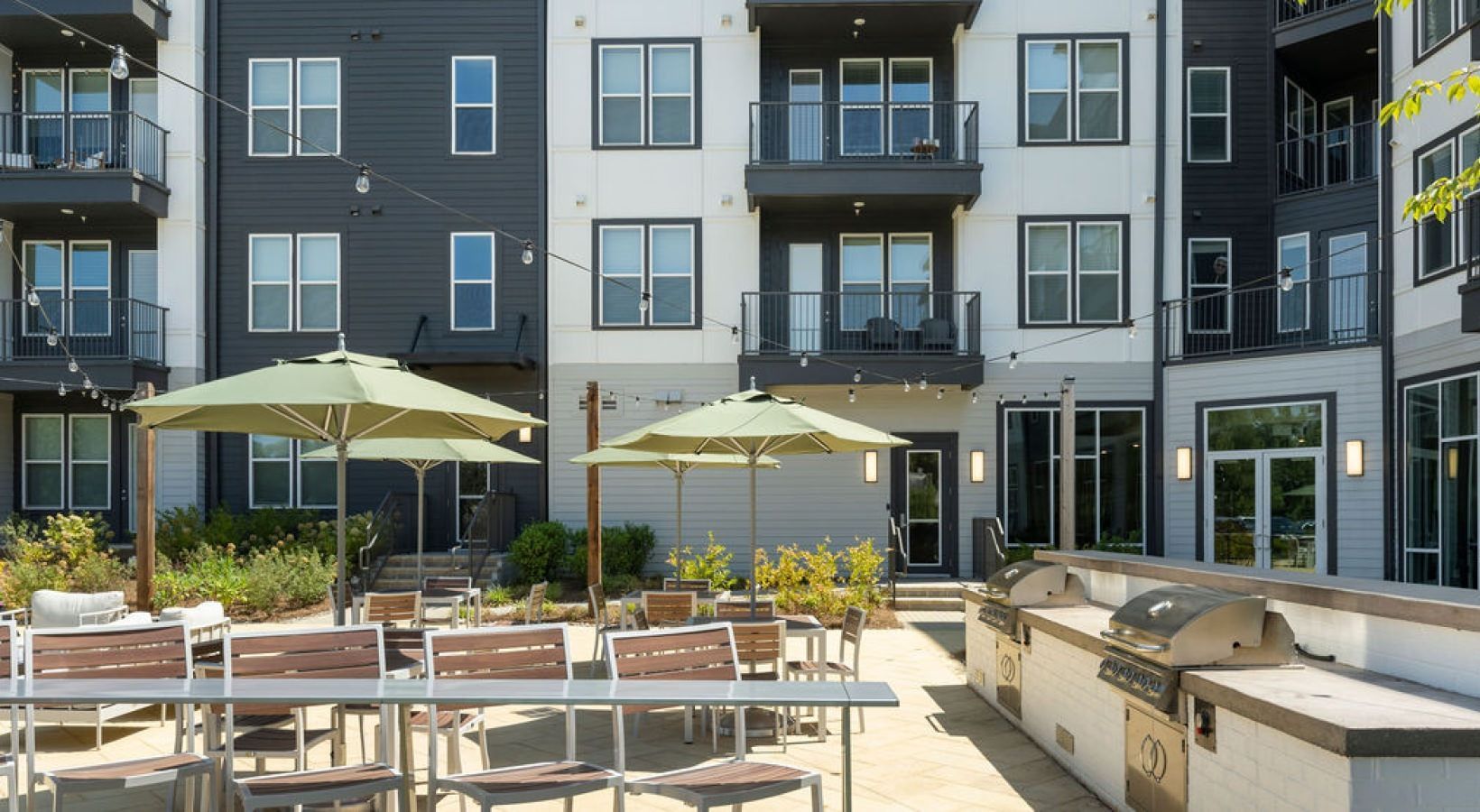 Outdoor communal courtyard at apartment complex with tables, umbrellas, grill, and string lights.