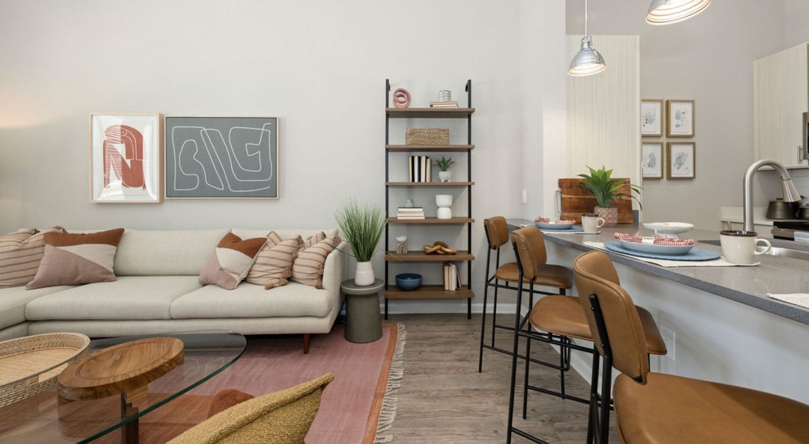 Open living area with beige sofa, wood-and-metal bookshelf, and a kitchen island with brown bar stools.