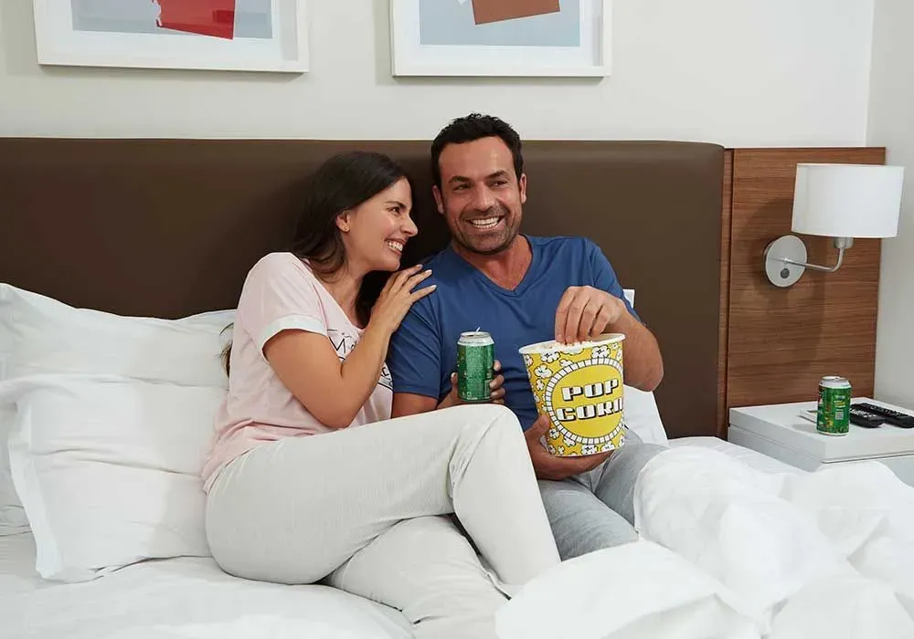 Um homem e uma mulher estão sentados em uma cama comendo pipoca e bebendo cerveja.