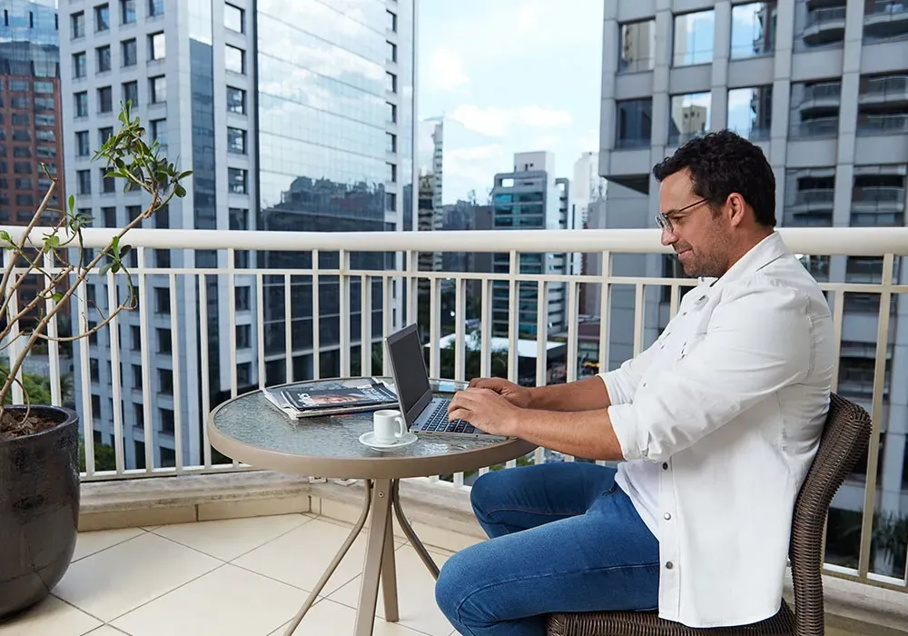Um homem está sentado em uma mesa na varanda usando um laptop.