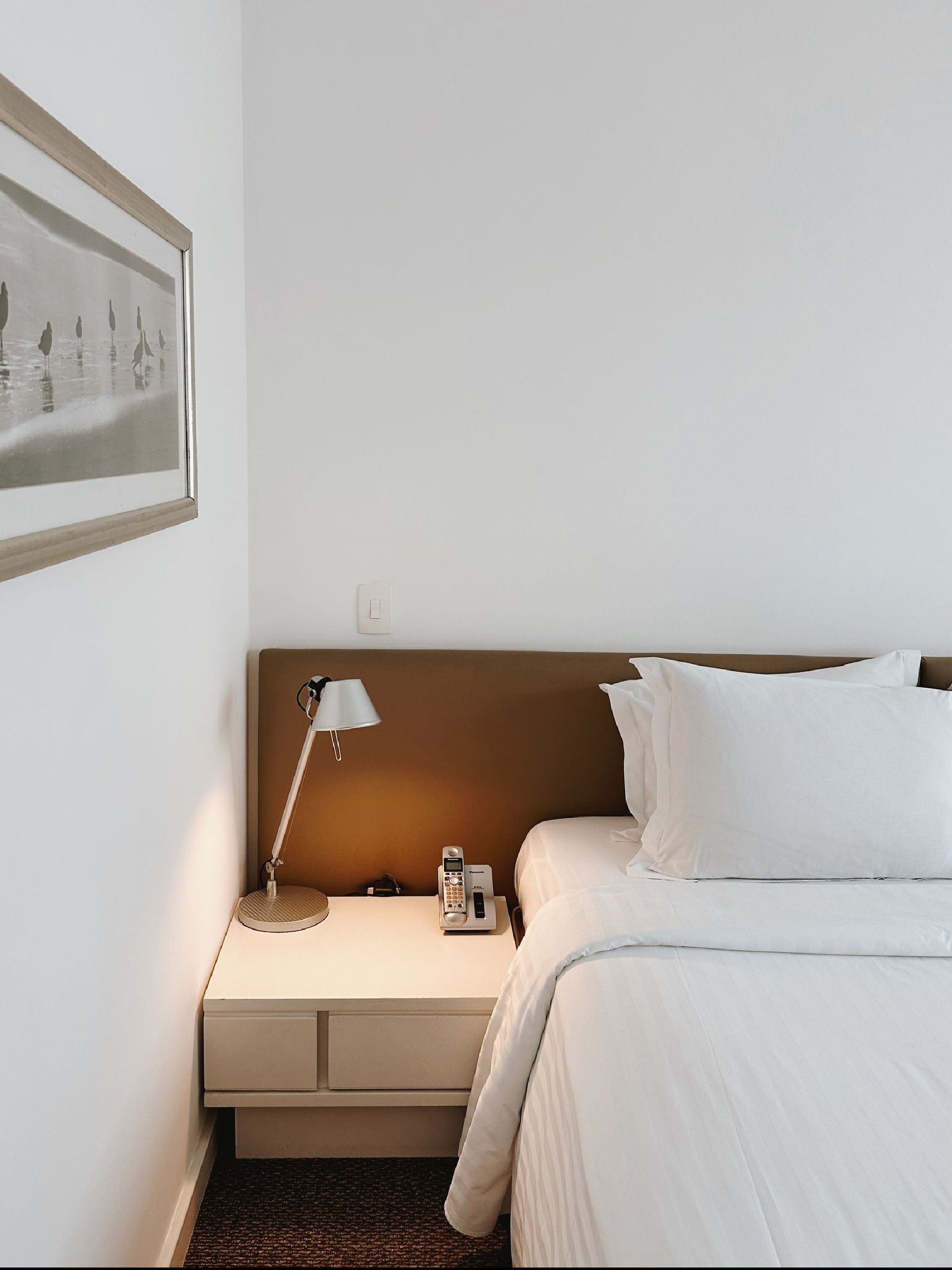 Um quarto com uma cama, mesa de cabeceira, abajur e um quadro na parede.