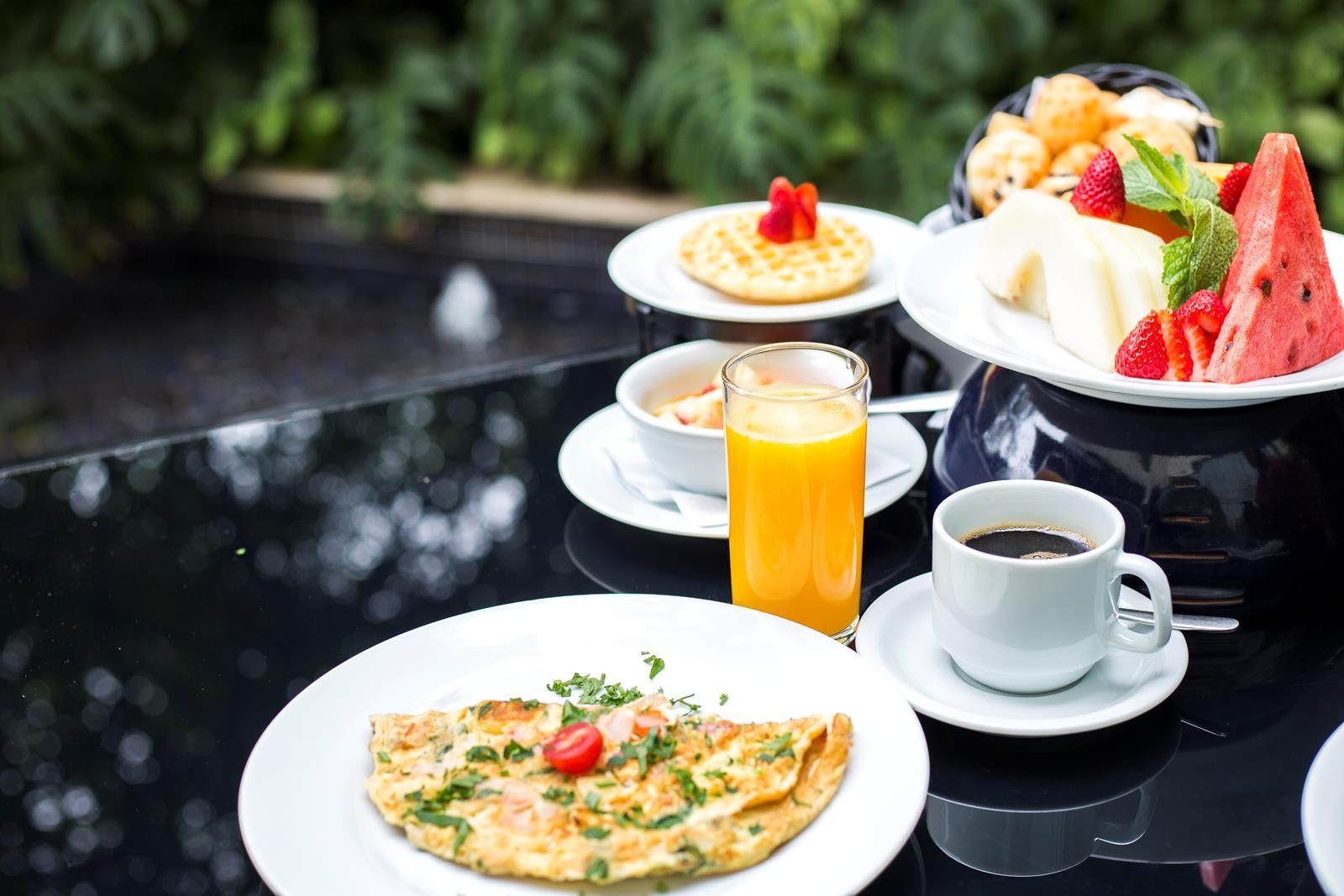 Café da manhã completo em área externa da eConfor Berrini, com omelete, frutas frescas, waffle, suco e café, destacando o serviço de hotelaria de alto padrão.