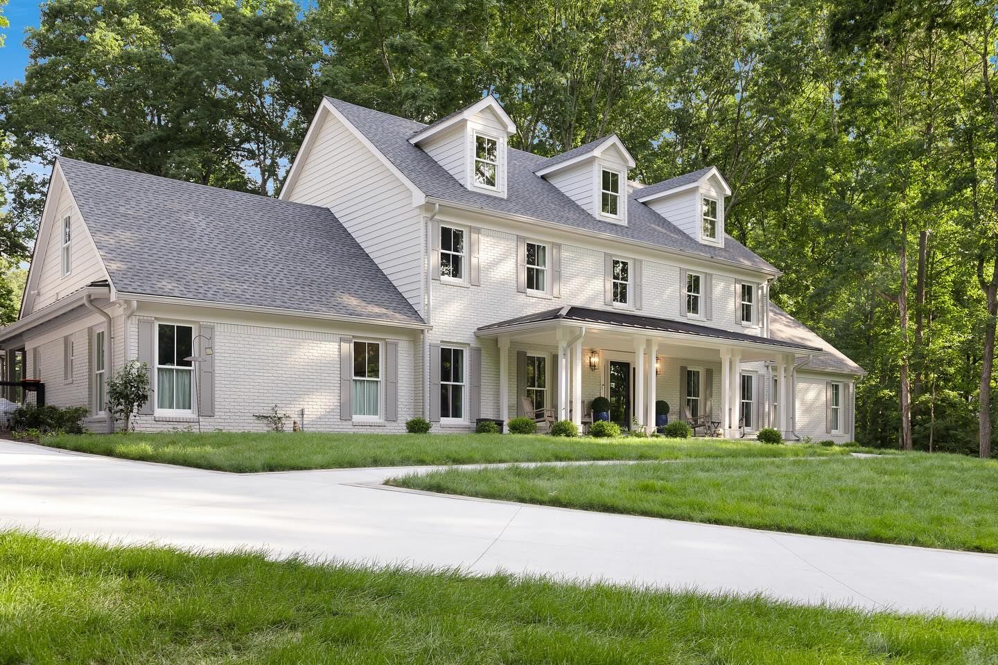 Two-story white farmhouse with gray roof, porch, and dormers, set on a grassy lawn with a winding driveway.