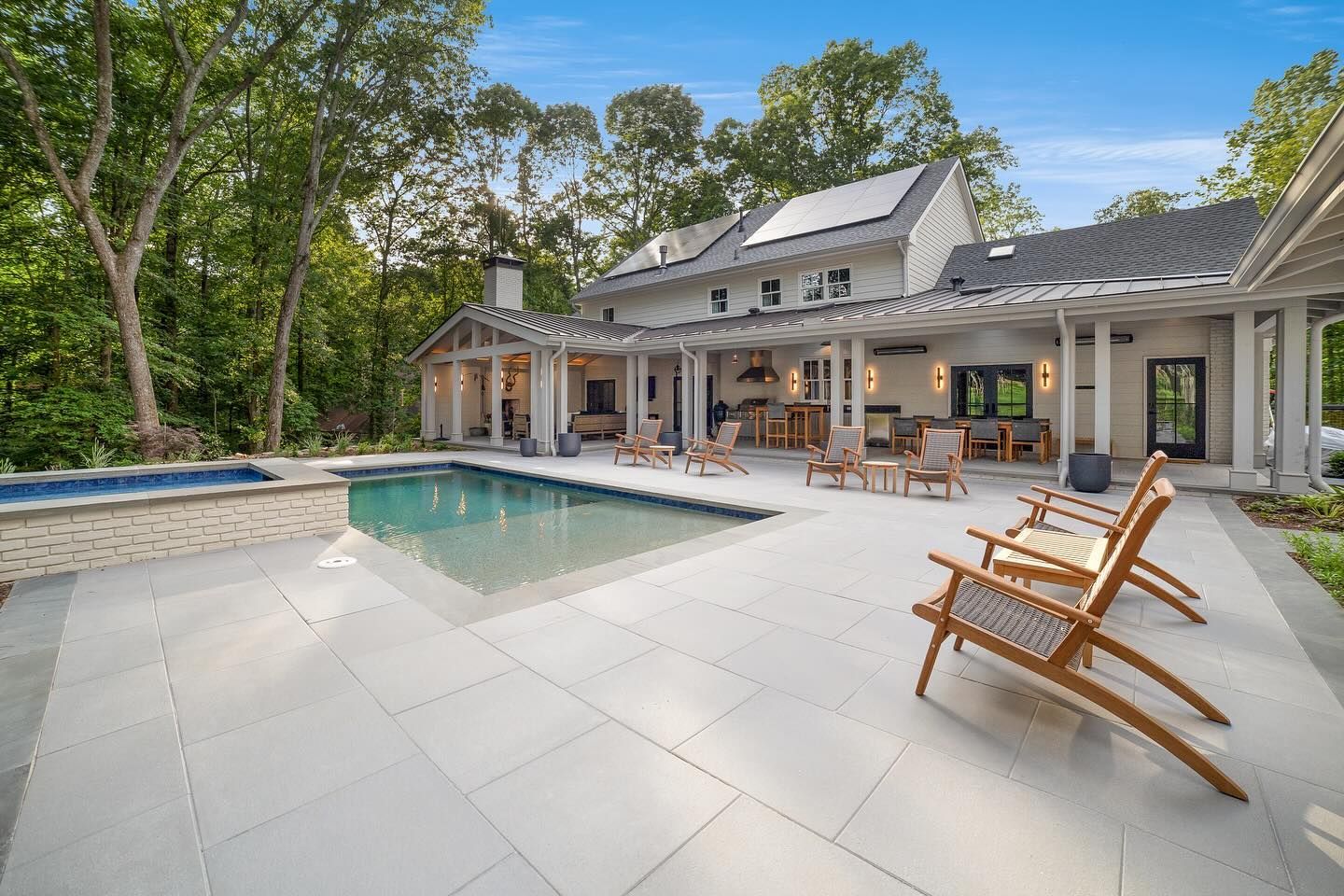 Backyard patio with a pool and lounge chairs; surrounded by a home and trees under a blue sky.