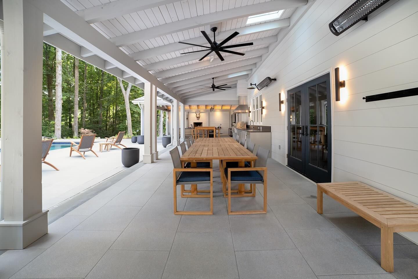Covered outdoor dining area with long table, chairs, kitchen, and pool view. White walls and ceiling, wood accents.