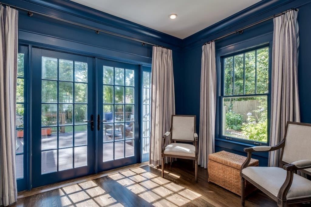 Blue-walled room with French doors, a window, two chairs, and light curtains, showing a sunlit outdoor view.