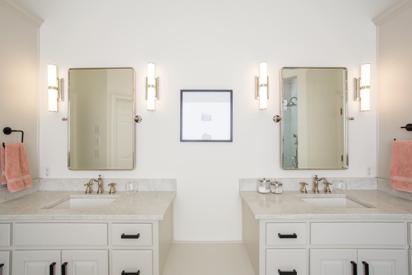 Bathroom with two white vanities, mirrors, and sconces. Pink towels and a framed artwork.