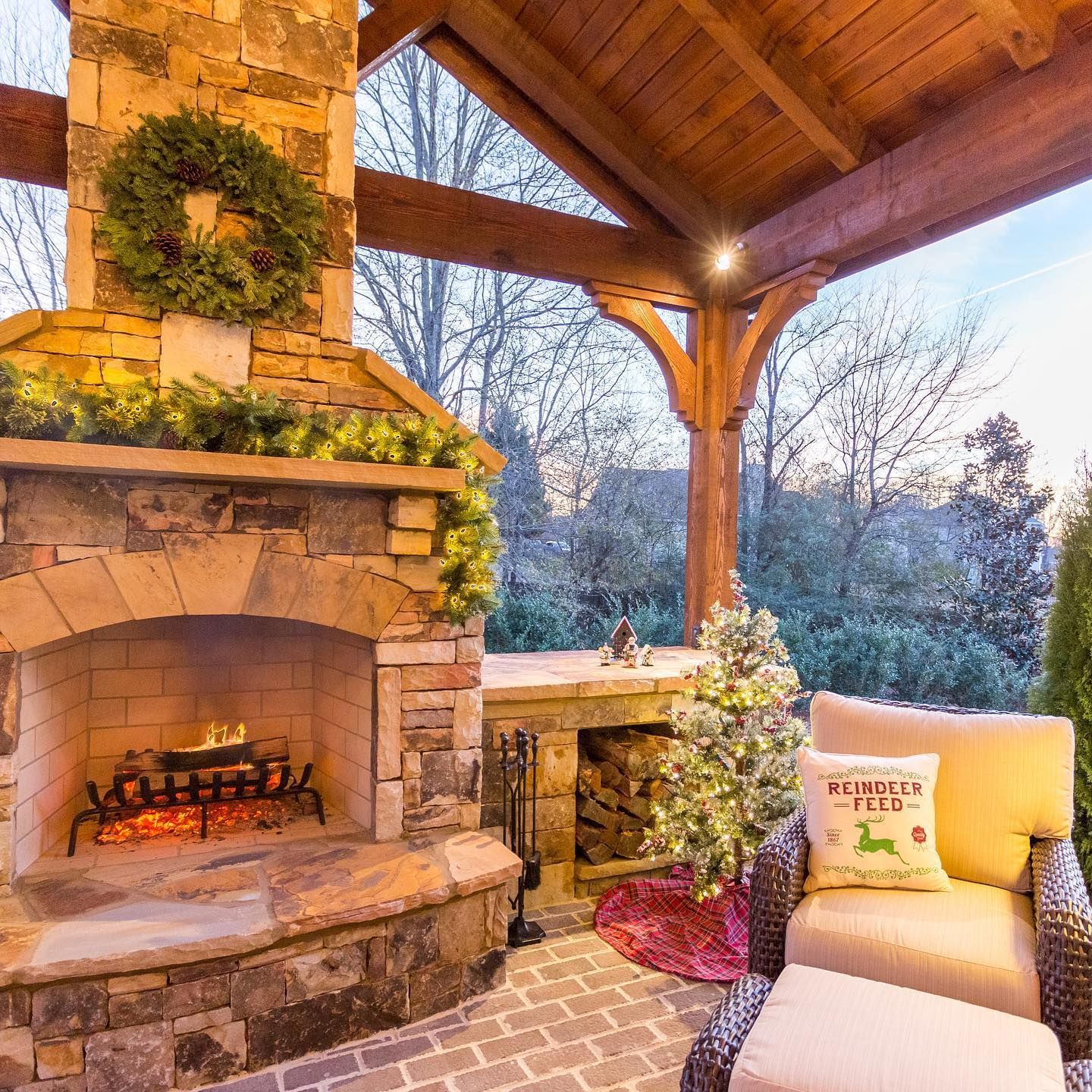 Outdoor fireplace with Christmas decor, under a wooden roof. Stone exterior, cozy seating.