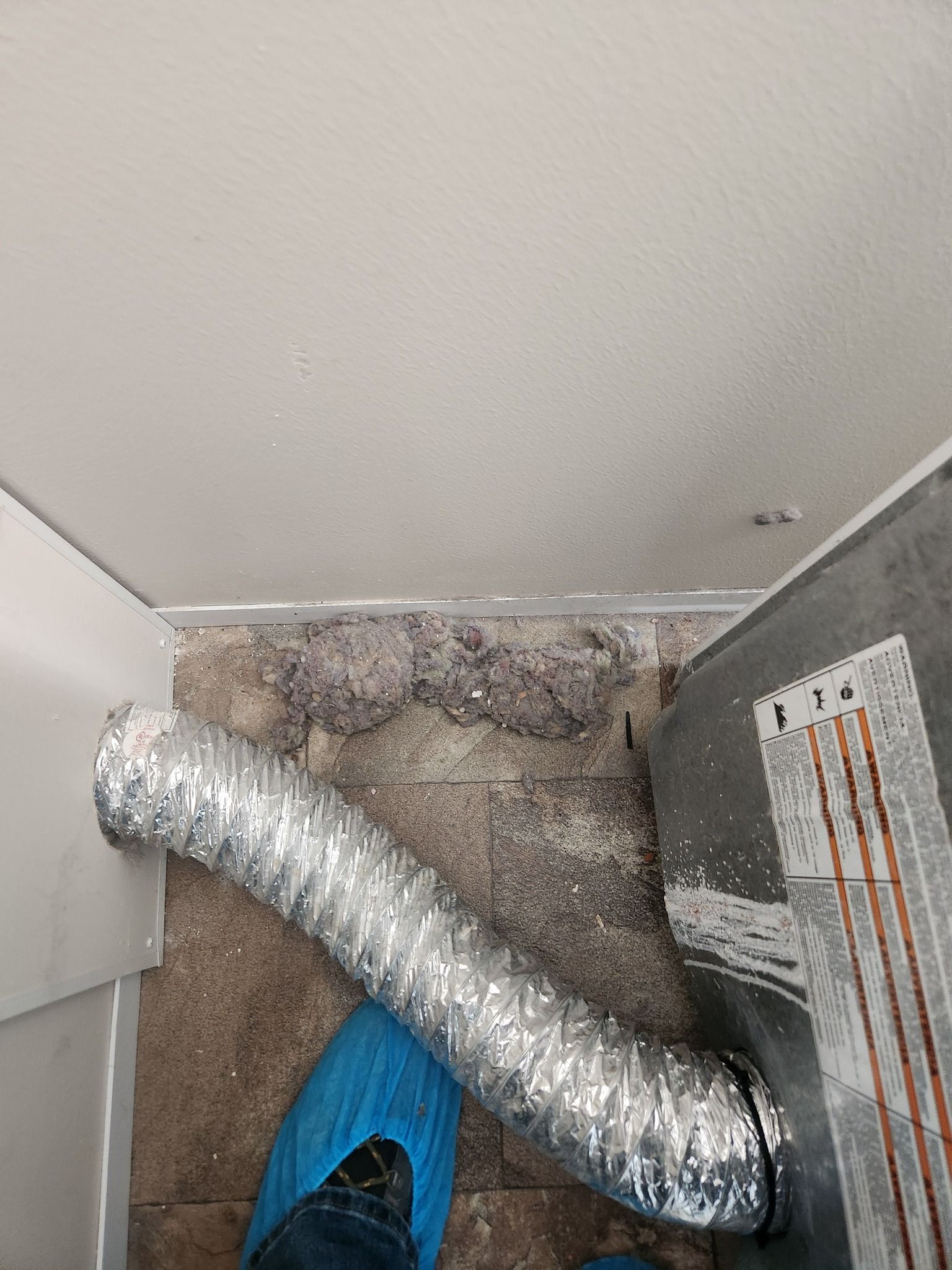 A dryer vent with lint buildup on the floor; blue shoe covers, and a white wall are also visible.