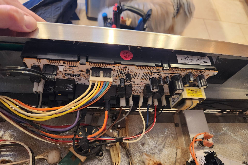 Close-up of an oven control board with colored wires and a hand holding the stainless steel trim.