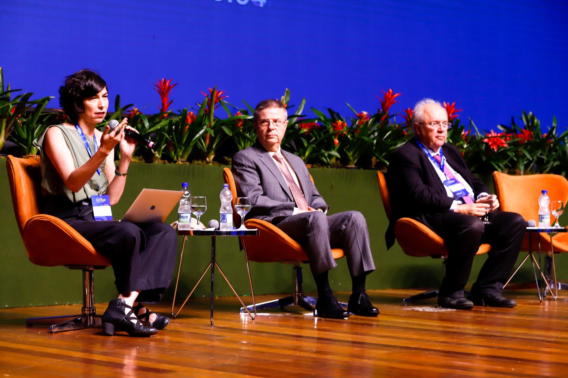 A woman is speaking into a microphone while two men sit in chairs.