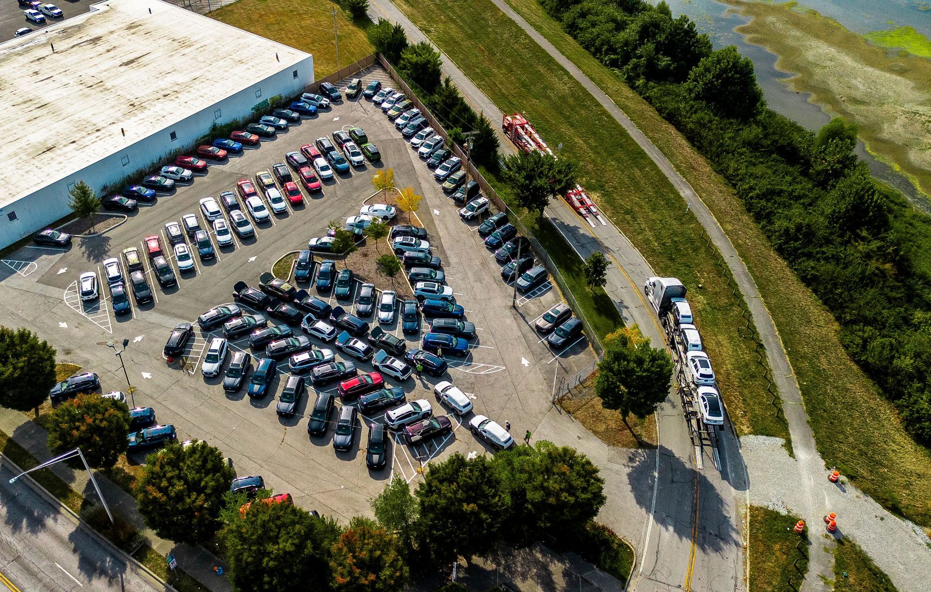 Aerial view of a car lot with many parked vehicles and a large building nearby; some cars are being loaded.