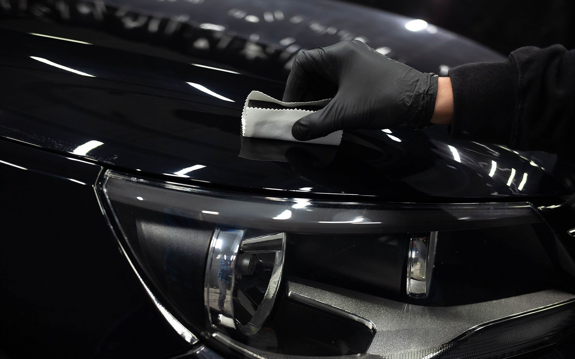 Black-gloved hand applying ceramic coating to a black car hood. Shiny surface, headlight visible.