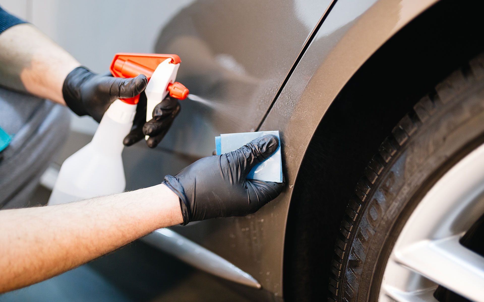 Person applying detailing product to car's side panel with a spray bottle and applicator, wearing black gloves.