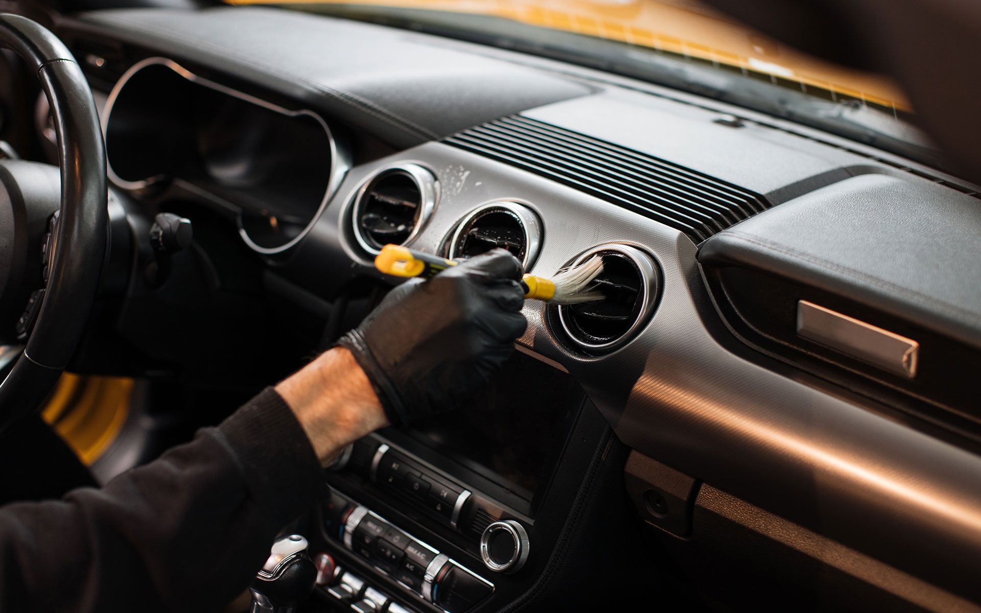 Gloved hand cleaning car air vent with a brush. Black dashboard with circular vents is visible.