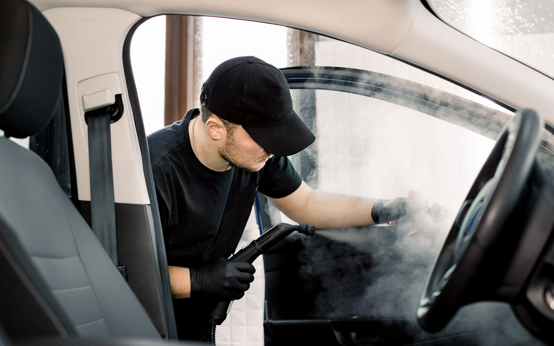 Man steaming car interior, cleaning door window. Black cap and gloves, steam visible.