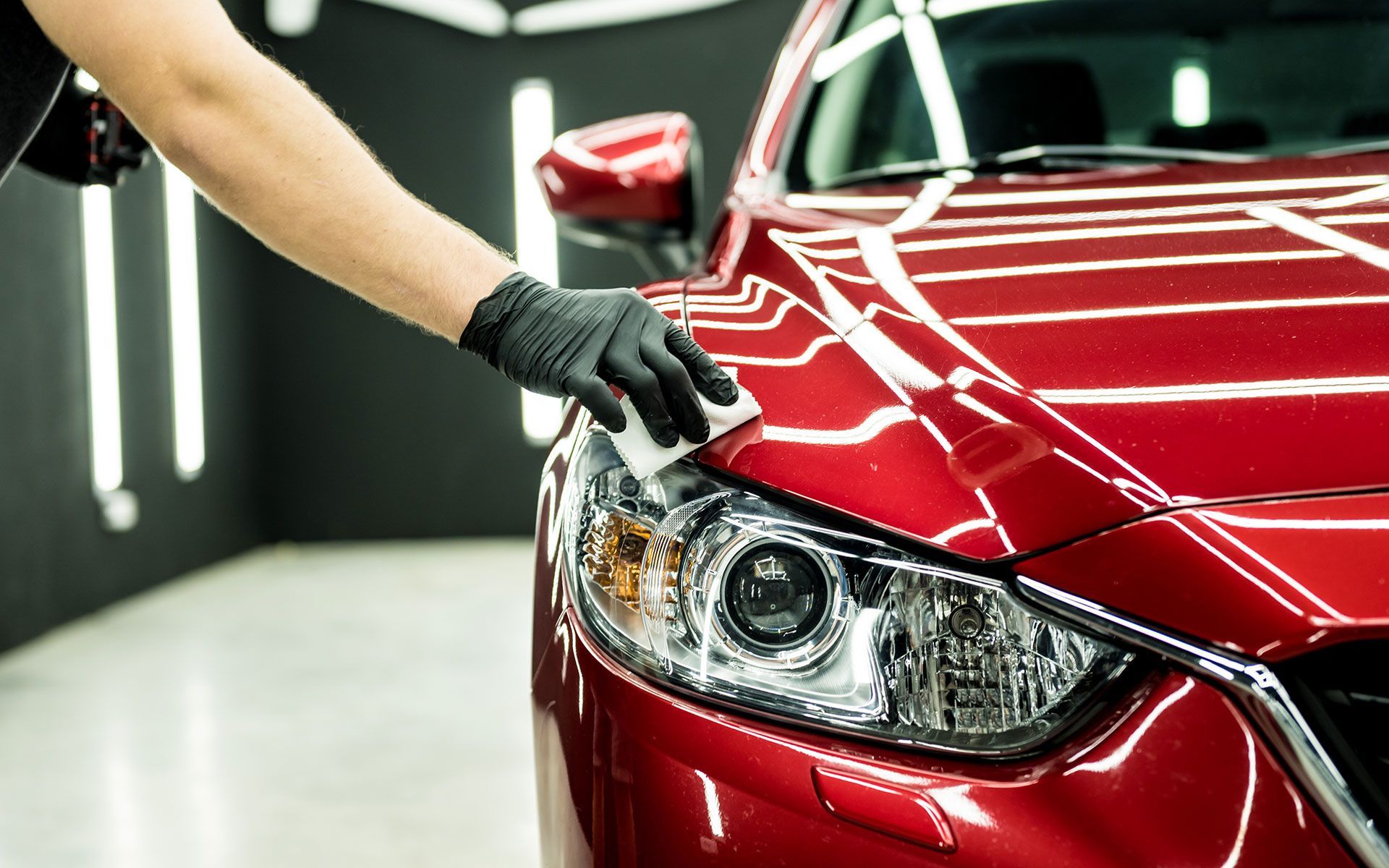 Gloved hand applying detailing product to a shiny red car's hood in a professional workshop setting.