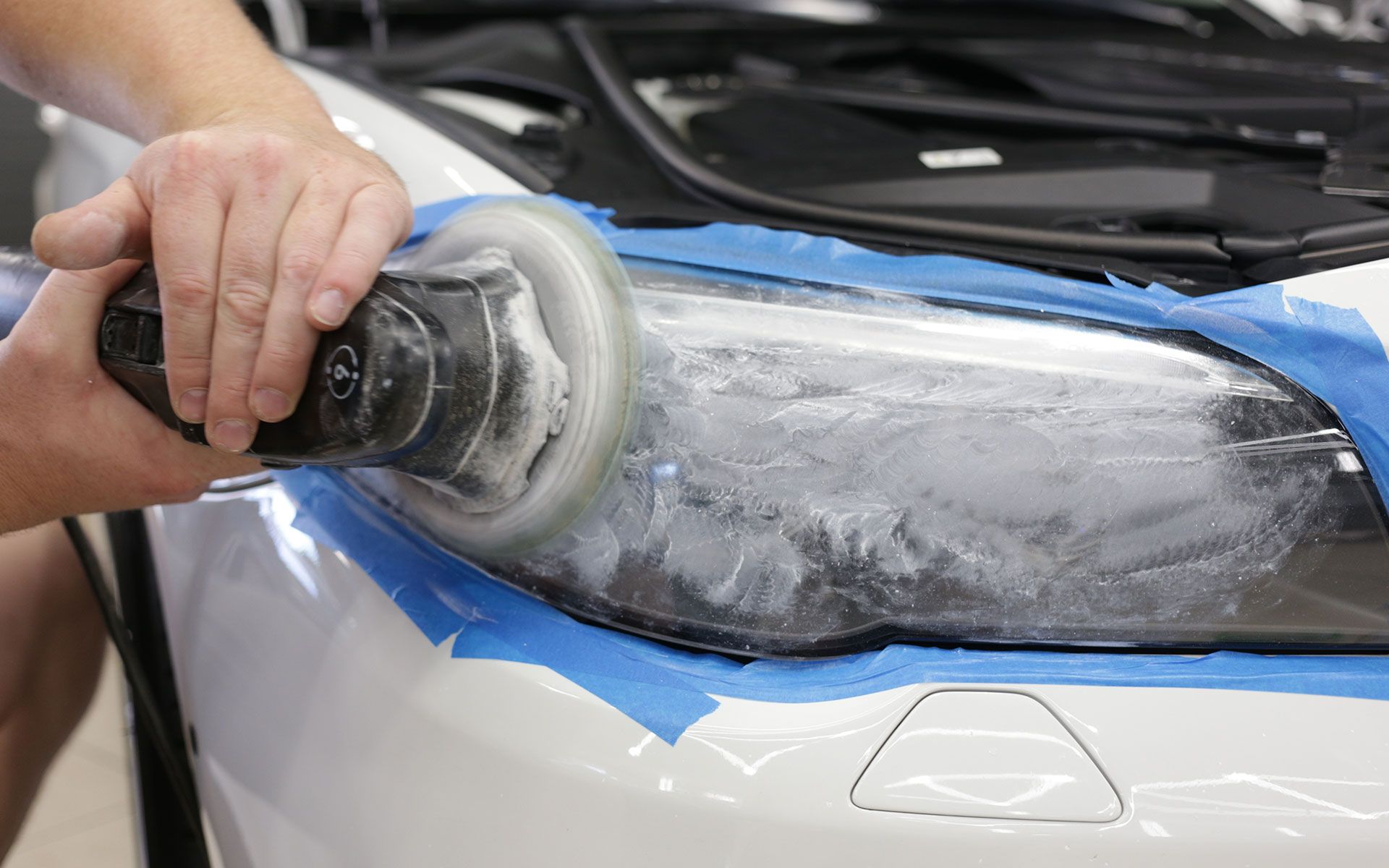 Person using a power buffer to polish a car headlight. Blue tape protects surrounding paint.