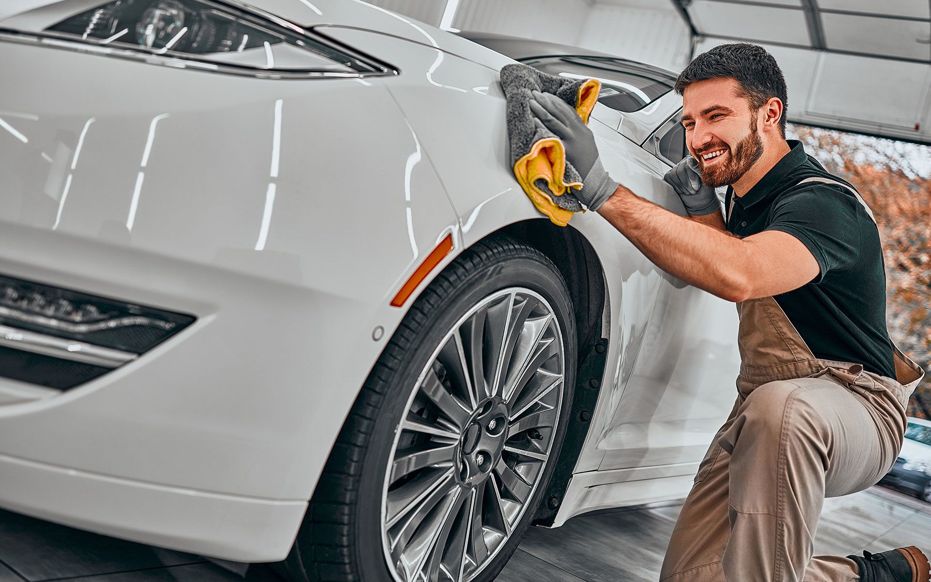 Man wiping down white car with yellow cloth; wearing gloves and smiling, outdoors.