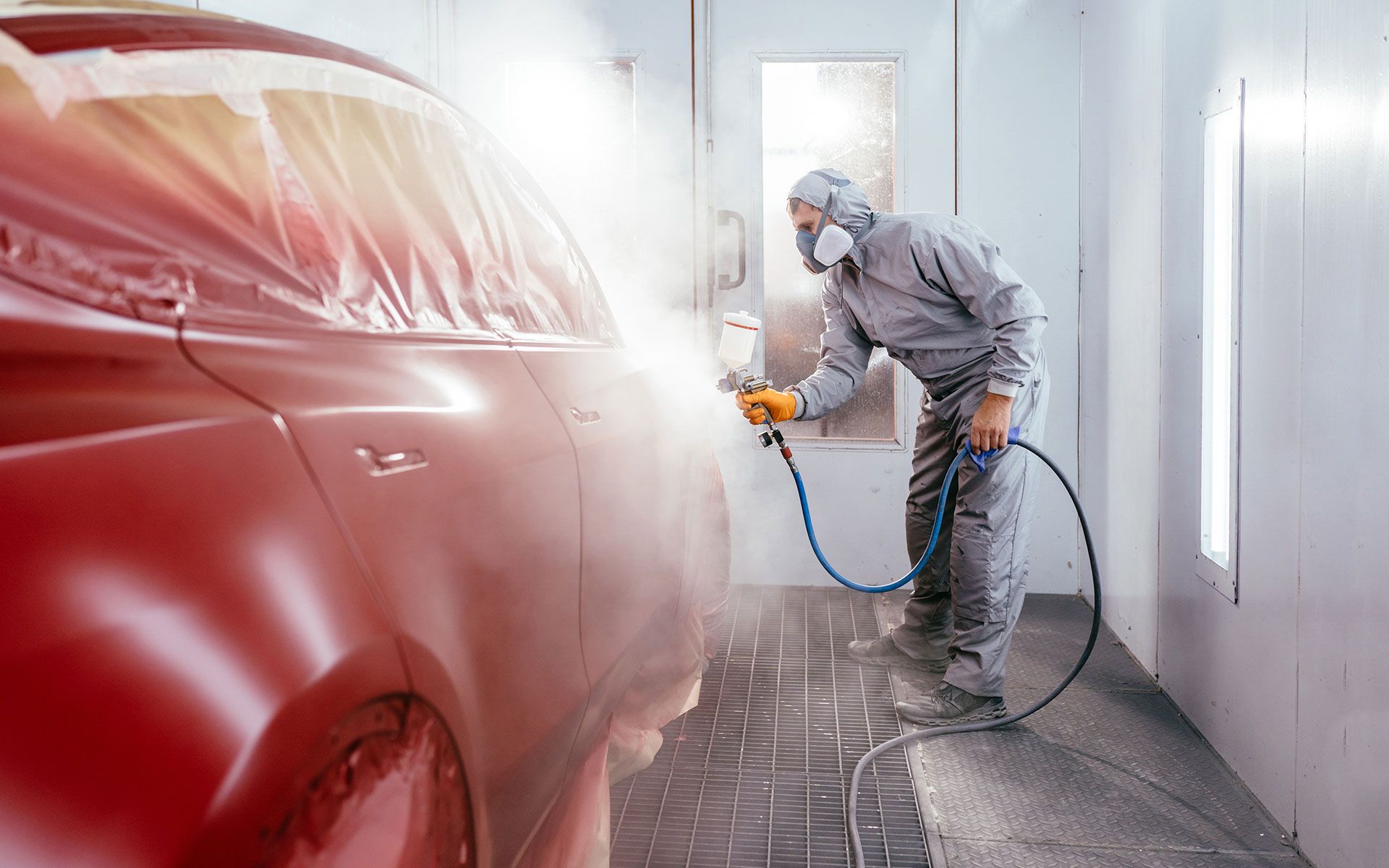 Person in protective suit spray-painting a red car in a paint booth.