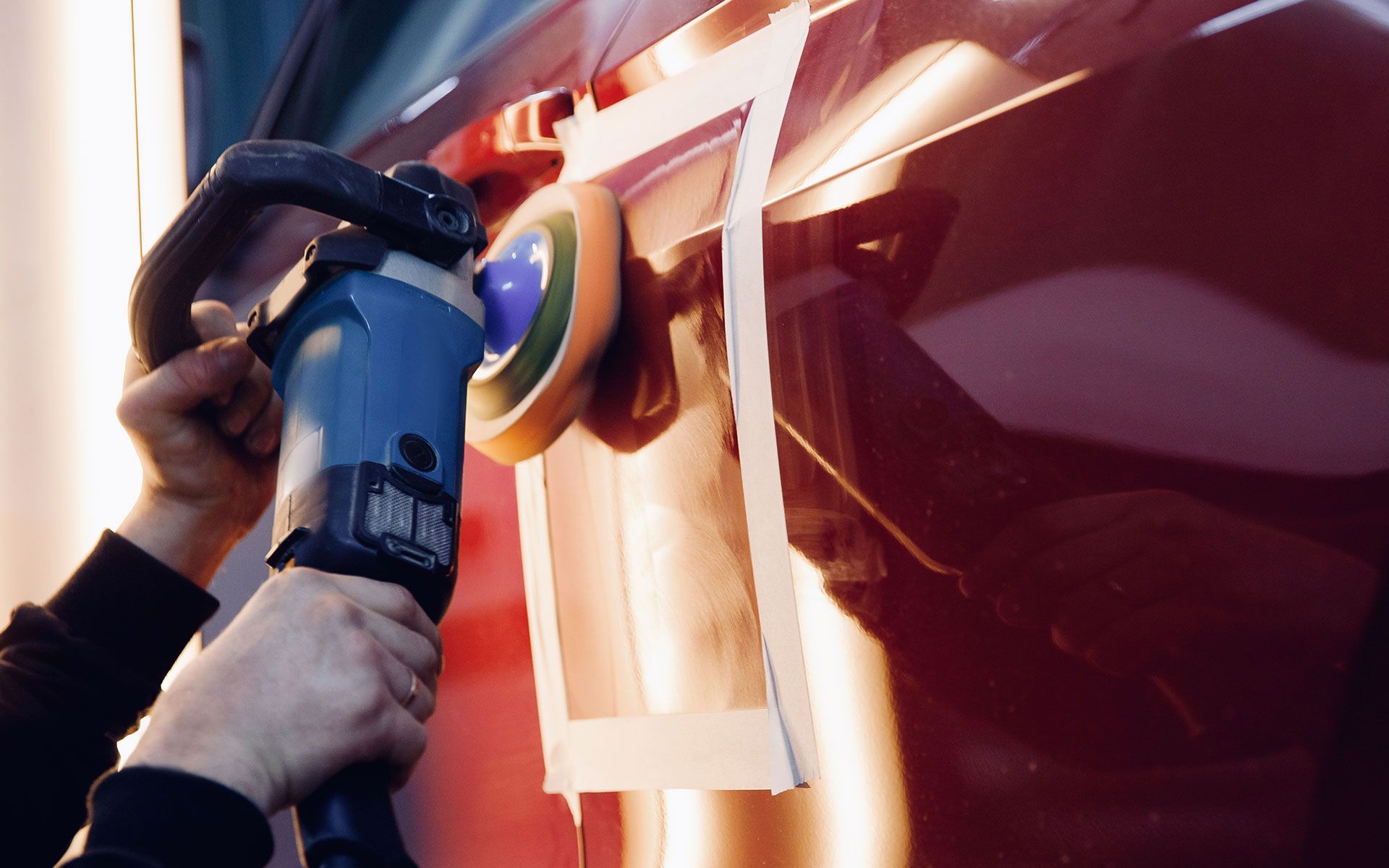 Person using an electric polisher on a red car door, with tape outlining the work area.