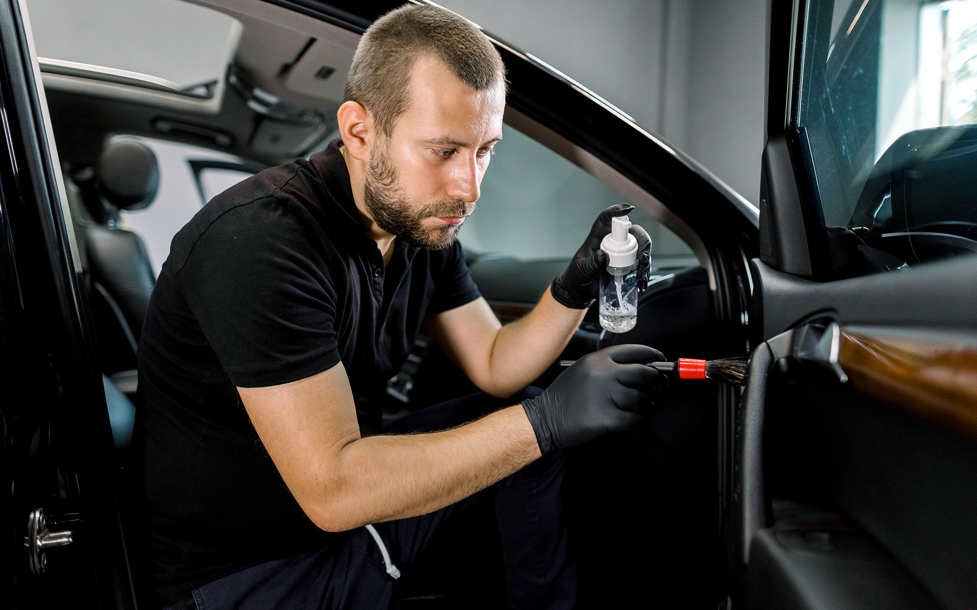 Man cleaning a car interior. He sprays a liquid and uses a brush on the door panel, wearing black gloves.