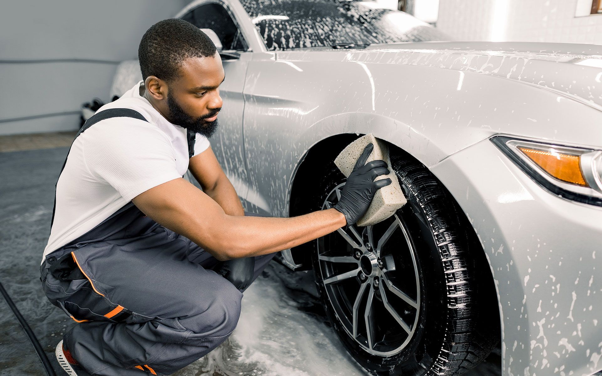 Man washes a silver car tire with a sponge, in a car wash bay.