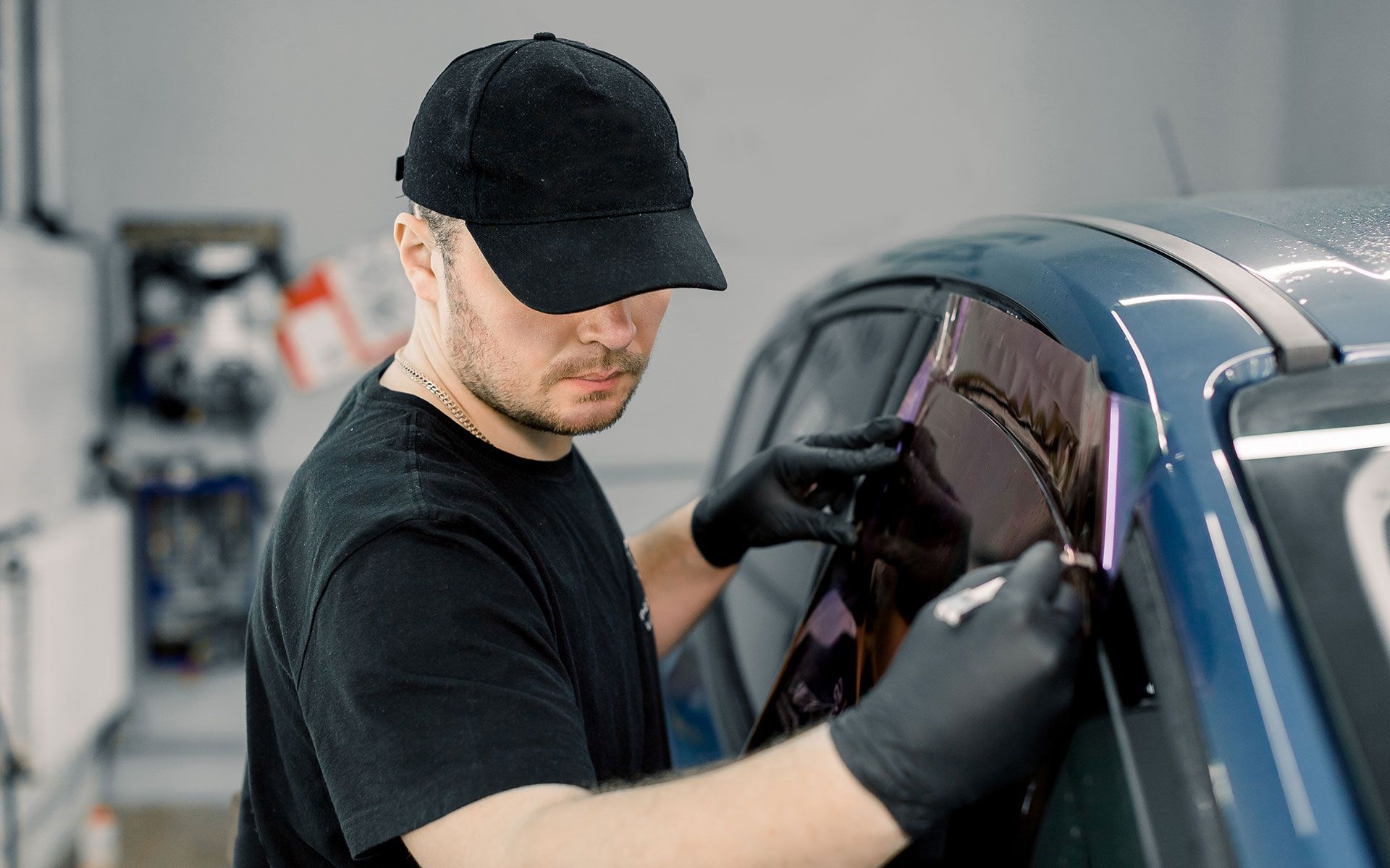 Person in black cap and gloves installing tinted window film on a blue car.