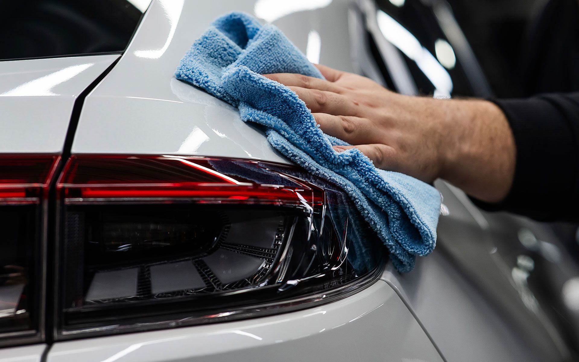 Hand wiping a blue microfiber cloth over a white car's taillight.