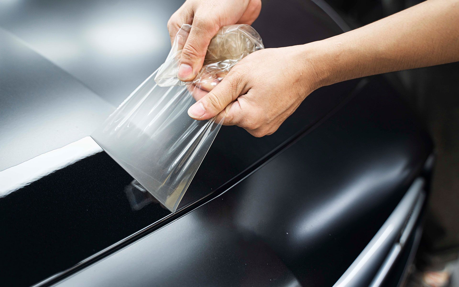 Hands peeling protective film from a dark car surface, likely paint protection film (PPF) application.